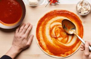 Close-up of a chef's hand spreading tomato paste on pizza dough